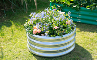 A round, galvanized metal raised garden bed showcasing a vibrant display of blue and peach flowers on a sunny lawn.