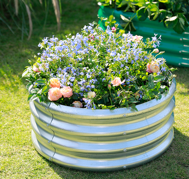 A round, galvanized metal raised garden bed showcasing a vibrant display of blue and peach flowers on a sunny lawn.