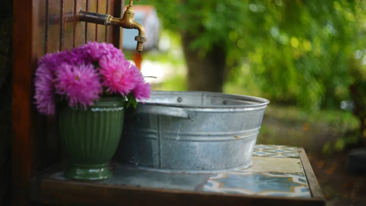 A galvanized tub sits on a tiled counter beneath an outdoor spigot, next to a green vase filled with pink flowers.