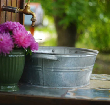 A galvanized tub sits on a tiled counter beneath an outdoor spigot, next to a green vase filled with pink flowers.