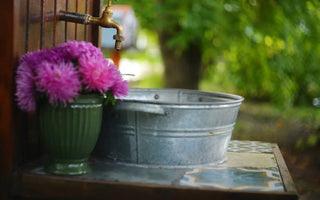 A galvanized tub sits on a tiled counter beneath an outdoor spigot, next to a green vase filled with pink flowers.