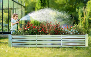 Child watering colorful flowers in a rectangular corrugated metal raised garden bed with greenhouse in background