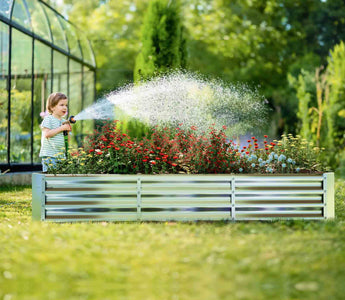 Child watering colorful flowers in a rectangular corrugated metal raised garden bed with greenhouse in background